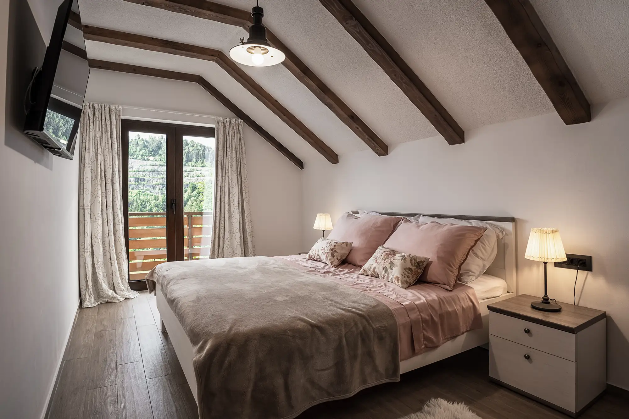 Potkrovna spavaća soba s posteljinom u ružičastim tonovima, drvenim gredama i pogledom na planine s balkona